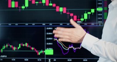 İş kıyafetleri analisti ticaretle ilgili bilgi verir. Erkek kişi plazma ekranında dinamik para birimi diyagramının arka planında el kol hareketleriyle duruyor