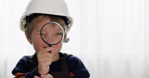 戴着白色头盔的有趣的学龄前儿童通过放大镜看相机 快乐的男孩扮演建设者的角色 带着郁金香在灯光下嬉笑 — 图库视频影像
