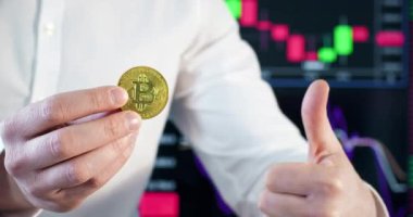 Hafif iş gömleği giyen tüccar Bitcoin 'i elinde tutuyor. Erkek kişi, ofiste ekranda dinamik diyagramın arka planında başparmak işareti gösteriyor.