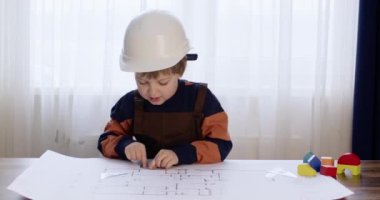 Beyaz kasklı küçük bir çocuk mühendis rolü oynuyor ve cetvelle ev çizimini ölçüyor. Çocuk daire tasarımı için yapı bloğunu hevesle seçti