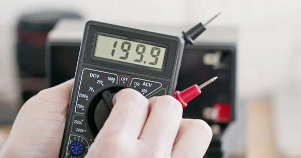 Man turns lever of multimeter and checks up accumulator battery in workshop close view. Technology of electrical equipment testing