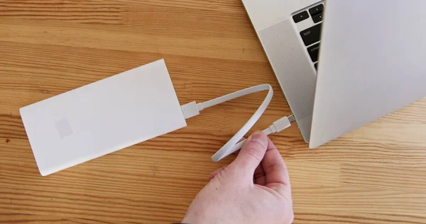 Person plugs power bank to laptop computer with cable on wooden table close upper view. Portable battery with large capacity for laptop charging