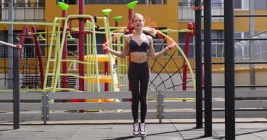 Spor giyimli genç bir kadın güneşli bir günde açık havada ip atlıyor. Kadın gündüz vakti şehir spor sahasında spor yapıyor. Sağlıklı yaşam tarzı yavaş çekim