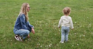 Genç anne park çayırındaki güzel çiçekleri yeni yürümeye başlayan çocuğa işaret ediyor. Oğlu, bahar şehir parkında yeşil çayırlarda karahindibalara doğru yürüyor.