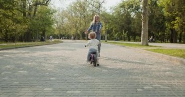 Genç anne, oğlunu şehir parkında mor dengeli bisiklete binerken yakaladı. Bisiklet süren çocuk, şehir parkındaki kaldırımda kadın ebeveyne yetişti.