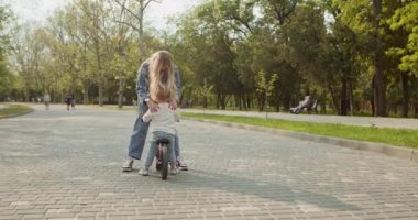 Genç anne, oğlunu güneşli şehir parkında bisiklete binerken yakaladı. Bisikletli çocuk şehir parkının kaldırımında kadın ebeveyne yetişiyor.