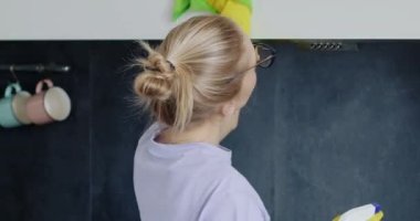 Sarışın kadın mutfak raflarını su spreyi ve bezle temizliyor. Yorgun yüzlü gözlüklü genç ev hanımı ev işlerini yapıyor.