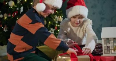 Anaokulu kardeşleri Noel hediyelerini süslü ağaçlara karşı açıyorlar. Noel Baba şapkalı abi ve abla yeni yılı kutluyor.