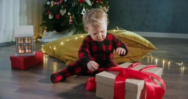 Küçük çocuk Noel arifesinde hediyeleri açmayı sever. Kırmızı pijamalı çocuk karton kutudan kırmızı kurdeleyi çıkardı ve yeni yıl hediyesini açtı.