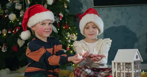 学龄前儿童手持玩具蜡烛 在圣诞树下装饰洋娃娃屋 圣诞礼帽上的兄弟姐妹们装饰着客厅过年 — 图库视频影像