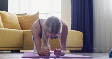 Gözlüklü sarışın kadın oturma odasında spor paspası üzerinde pilates yapıyor. Geniş bir dairede tahta egzersizleri yapan bir kadın.