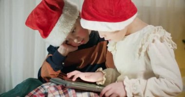 Evde kalan anaokulu öğrencileri favori çizgi filmleri izlemek için heyecan verici bir şekilde tablet kullanıyorlar. Noel Baba şapkası takan kardeşler tablette canlı yayın yapıyor.