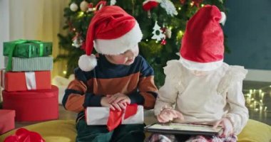 Anaokulu çocuğu ve kız süslü oturma odasında çizgi film izlemek için tablet aldı. Noel Baba şapkalı kardeşler hediyelere karşı birlikte çizgi film izlerler.