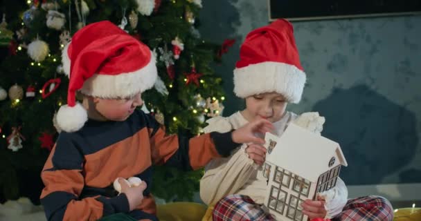学龄前儿童在圣诞树下用蜡烛玩具装饰洋娃娃屋 戴着圣诞礼帽的男孩和女孩装饰客厅 — 图库视频影像