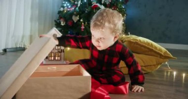 Bebek, Noel hediyelerini Noel ağacına karşı oturma odasında çelenklerle açıyor. Bebek apartmanda hediyelere bakmayı sever.