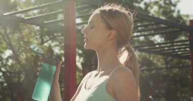 Spor giyimli genç bir kadın açık hava antrenmanından sonra ferahlatıcı bir yudum su alıyor. Kadın sporcu spor programını bir şişe tatlı suyla tamamladı.