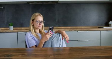 Genç sarışın kadın masada oturuyor ve cep telefonundan mesajlar okuyor. Dijital cihazlı eğlence modern teknolojiler evde mutfakta kullanılıyor
