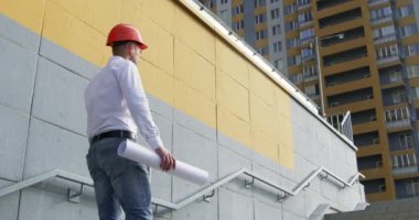 Miğferli mühendis, modern çok katlı binanın merdivenlerinde çizim yaptığını okuyor. Planlı müfettiş şehirdeki yeni apartman dairesini kontrol etti.