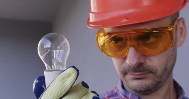 持怀疑态度的男电工穿着护目镜 看着灰色背景特写上的灯泡 技术员确保灯泡和灯都能正常工作 — 图库视频影像