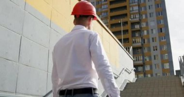 Yeni inşa edilen binanın yanındaki basamaklarda miğfer incelemesi yapan mimar. Elinde çizim olan mühendis şehirdeki yeni evi teftiş ediyor.