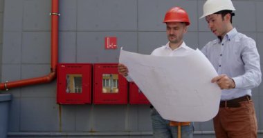 Erkek mimarlar gerçek binalarla karşılaştırıldığında plandan bahsederler. Miğferli müfettişler kağıtları gözden geçirip doğruluk hakkında konuşuyorlar..