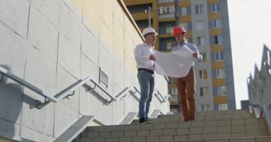 İnşaat alanı inşaatını inceleyen erkek mühendisler. Profesyonel müteahhitler basamaklarda duran binaya işaret eden mimari çizimleri tartıştılar