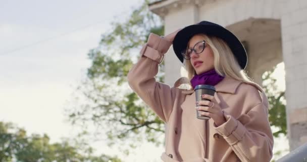 帽子とメガネをかぶった女性は コーヒーを飲む市内中心部の階段を下って行きます 曇りの日を明るくするために味付けされた飲み物と秋の散歩 — ストック動画