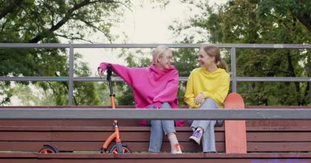 ティーンエイジャーの女の子は トリビューンベンチに座って歩いている人々について話します 女性の親友は 近くのスクーターとスケートボードを保持するコミュニケーションを楽しむ — ストック動画