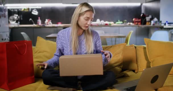 好奇心旺盛な金髪の女性はソファに座っているオンラインストアから受け取ったボール紙を開きます 女性のお客様がボックスに入り込むことに興奮する — ストック動画