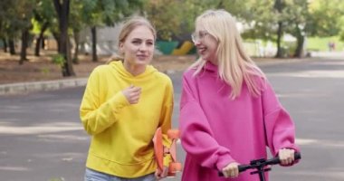 Genç kızlar scooter ve kaykayla yürürken gülerler. Sarışın gençler hafta sonu ağır çekimde kaykay parkına giderken şaka yaparlar.