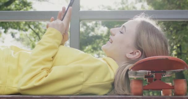 笑顔の女の子はスマートフォンでスケートボードのチャットで頭の上に横たわっています ハッピーティーンエイジャーはアクティブなトレーニング休憩中にベンチで休んでいる間 携帯電話を使用しています — ストック動画