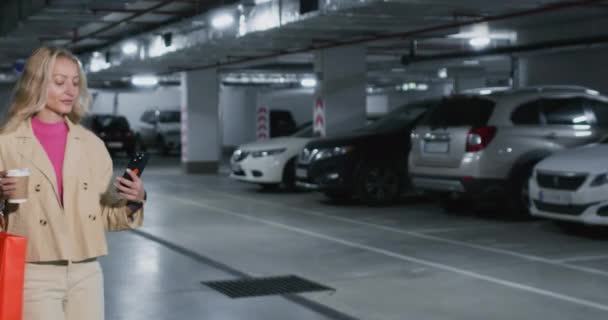 Stylowa Blondynka Spaceruje Podziemnym Parkingu Samochodami Kawą Torbami Zakupy Uśmiechnięta — Wideo stockowe