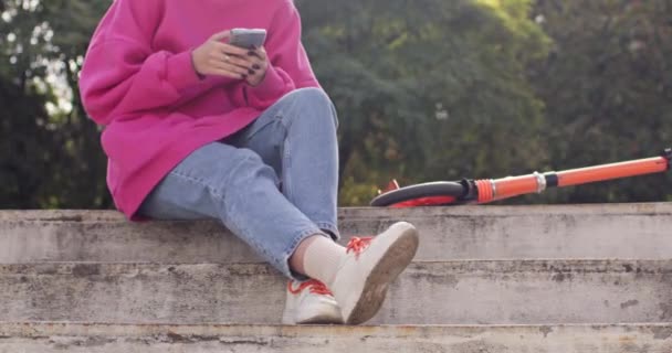 女の子はスクーターが付いているコンクリートの障壁に座っているスマートフォンを使用します ティーンエイジャーは友人にメッセージを入力するために 極端なライディングレッスンの間に休憩を取ります — ストック動画