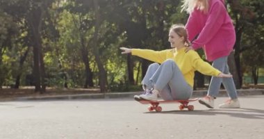 Kız kaykayla uçağın taklidini yaparken sarışın arkadaşı ona karşılık veriyor. Şehir parkındaki en iyi arkadaşının yardımıyla kaykay yapan genç bir kız.