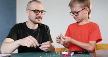 Mutlu baba, oğluna sofradaki inşaatçıların metal detaylarını vidalamayı öğretiyor. Gözlüklü çocuk ve adam evde modelleri bir araya getiriyorlar.