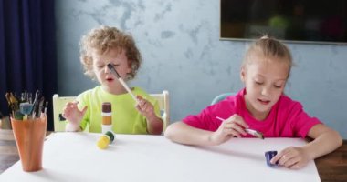 Kız fırçayı mavi boyaya batırıyor. Kız, boyayı çıkarmak için boya fırçasını kabın kenarına vuruyor. Çocuk kardeşler başyapıt yaratmaya hazırlanıyorlar.