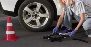 Lastik eldiven takan kadın kriko takmak için arabanın arka tekerleğine çömeliyor. Sarışın şoför yerde yatan makas krikosu alıyor. Trafik konisi yakınlarda duruyor.