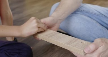 Anaokuluyla baba komodinin tahta parçalarını vidayla birleştiriyor. Erkek eller yonga tahtasını tutarken erkek çekiçler çivi çakıyor. Çocuklu bir adam yerde oturuyor.