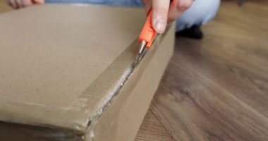 Yerde oturan adam karton kapağı et bıçağıyla açıyor. Lotus poziyonunda konumlandırılan kişi, kutunun kenarlarını keserek kümeyi göstermek için araç kullanır