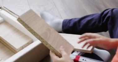 Anaokulu öğrencisi komodinin montajı için açık kutudan parçalar alıyor. Genç çocuk dikkatlice yonga tahtasını alıyor ve parça parça deliklerden bakıyor.