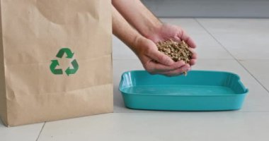 Adam ellerinden az miktarda kedi dolgusunu tepsiye döküyor. Sahibi, kum tanelerini hayvan kutusuna atarak kağıt torbadan ağır ağır toplar.