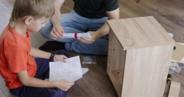 Erkek ve anaokulu öğrencisi yerde oturup ahşap komodinleri birleştiriyorlar. Küçük çocuk çizim yaparken baba parçaları birleştirmeyi anlatıyor.