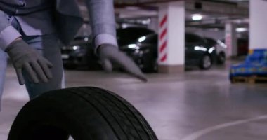 Adam yeraltı garajında arabanın önünde tekerlek lastiği yuvarlıyor. Lastik değiştirme işlemi sırasında eldivenli sürücü elementleri zemin boyunca hareket ettirir