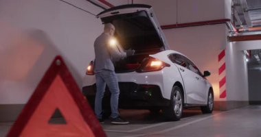 Eldivenli sürücü trafik işaretinin arkasında araba lastiklerini değiştiriyor. Adam bagajı tehlikeli uyarı ışıklarıyla açıyor lastikleri değiştiriyor.