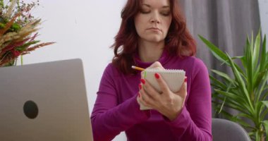 Esmer girişimci değerlendirmeleri dizüstü bilgisayarda çalışır ve not defterine not alır. Kadın, işin en önemli detaylarını küçük not defterine hızlı yazıyor.