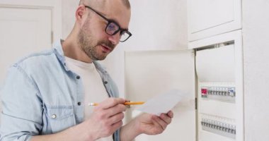 Gözlüklü adam güç paneli operasyonunu anlamaya çalışıyor. Kafası karışmış kel adam talimatları okur ve elinde kalemle düğmeleri çevirir.