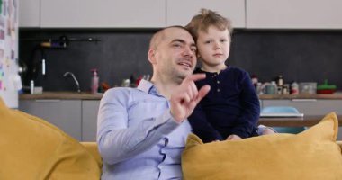 Küçük çocuk babasıyla yumuşak koltukta dinleniyor. Sarışın, sevimli çocuk mutlu babaya sarılıyor ve stüdyo dairesindeki minderlerin üzerinde dans etmeye başlıyor.