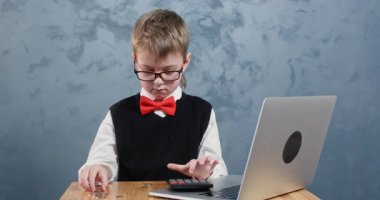 Okul üniformalı çocuk bilgisayarın yanındaki bozuk paraları hesaplıyor. Gözlüklü odaklı öğrenci masasında toplamda kaç para kazanacağını hesaplıyor.