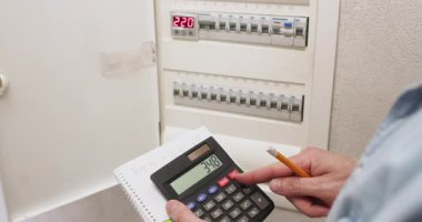 Adam devre kesici panelden ölçümler yazıyor. İşçi hesap makinesindeki verileri hesaplar ve problemi çözmek için elinde kalemle anahtarları çevirir