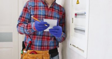 Erkek elektrikçi elektrik şalteri paneli hakkında notlar alıyor. Mavi eldivenli işçiler duvara yaslanırken bilgilerini not ederler.